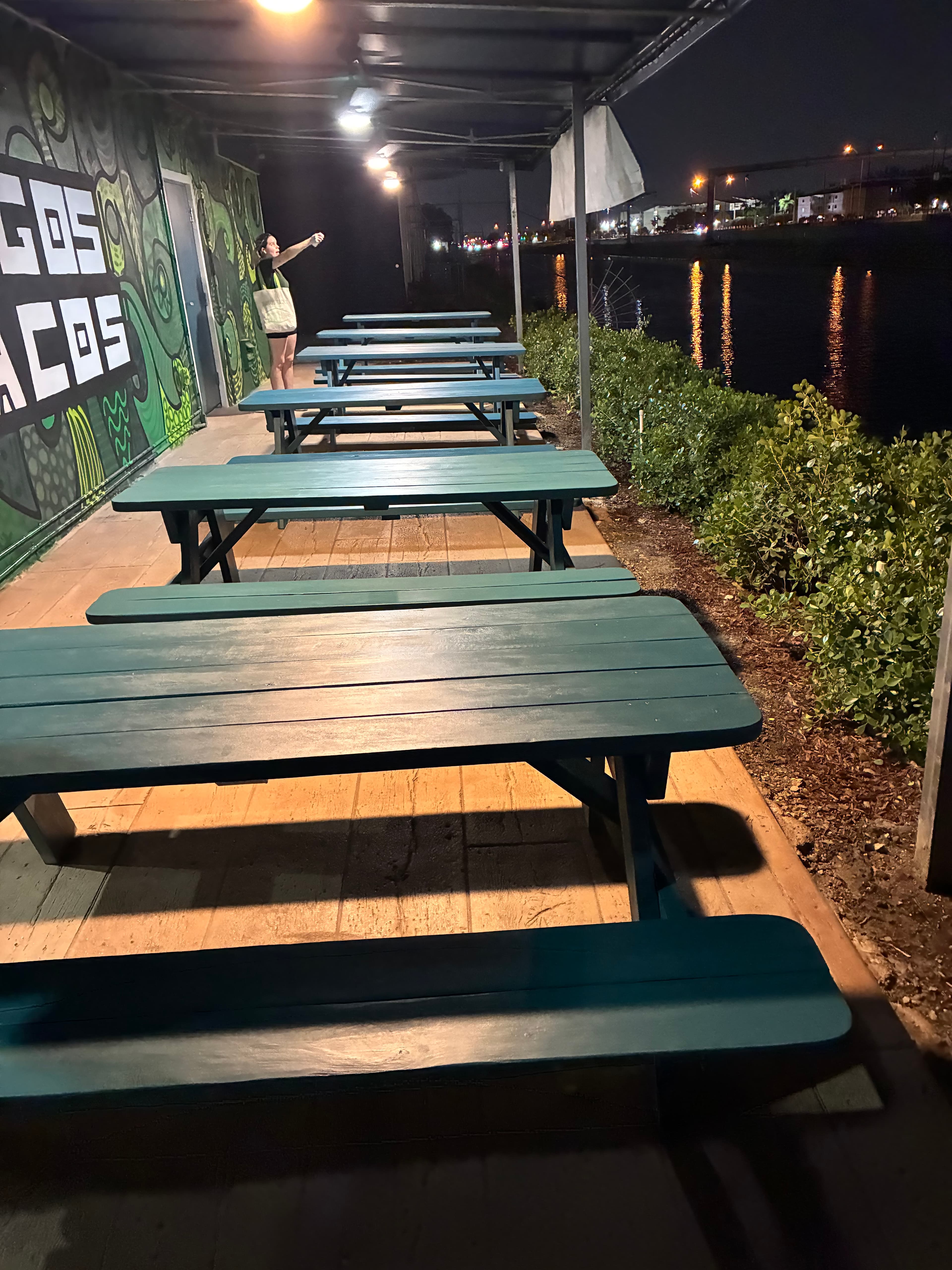Teal picnic tables on a night patio beside a river with glowing city light reflections.