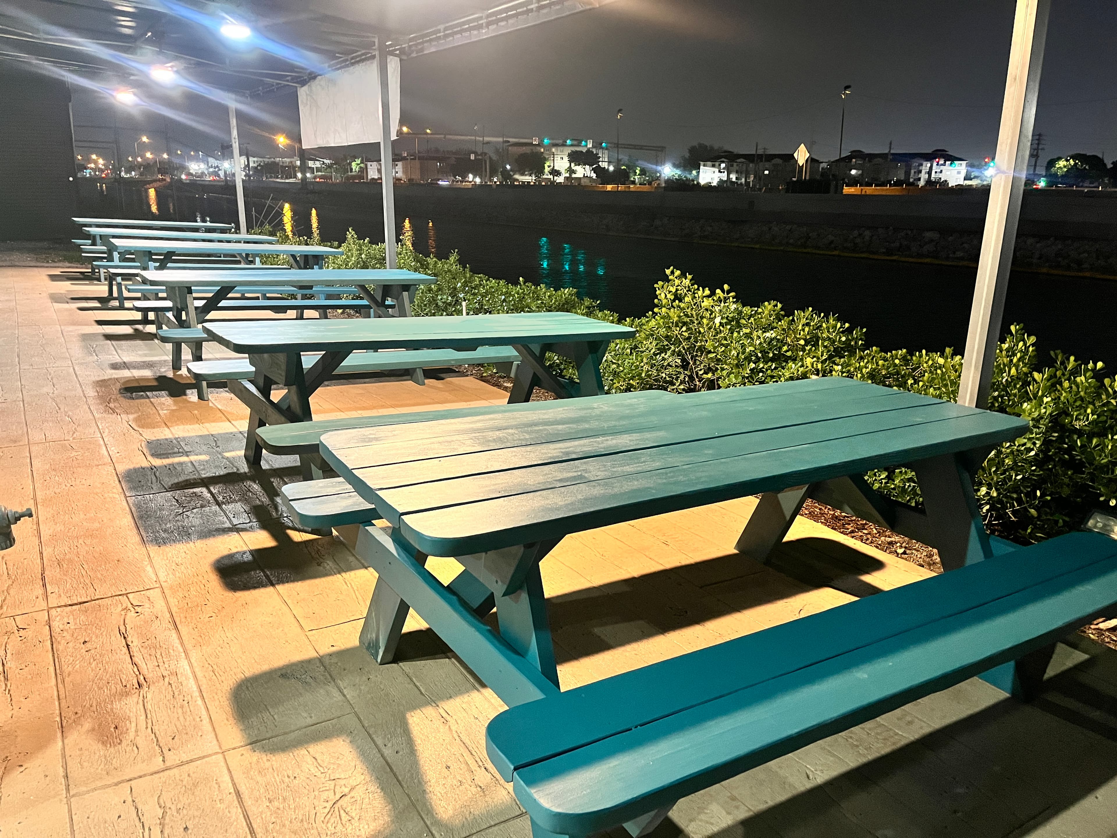 Teal picnic tables lined up on a patio at night overlooking a dark canal.